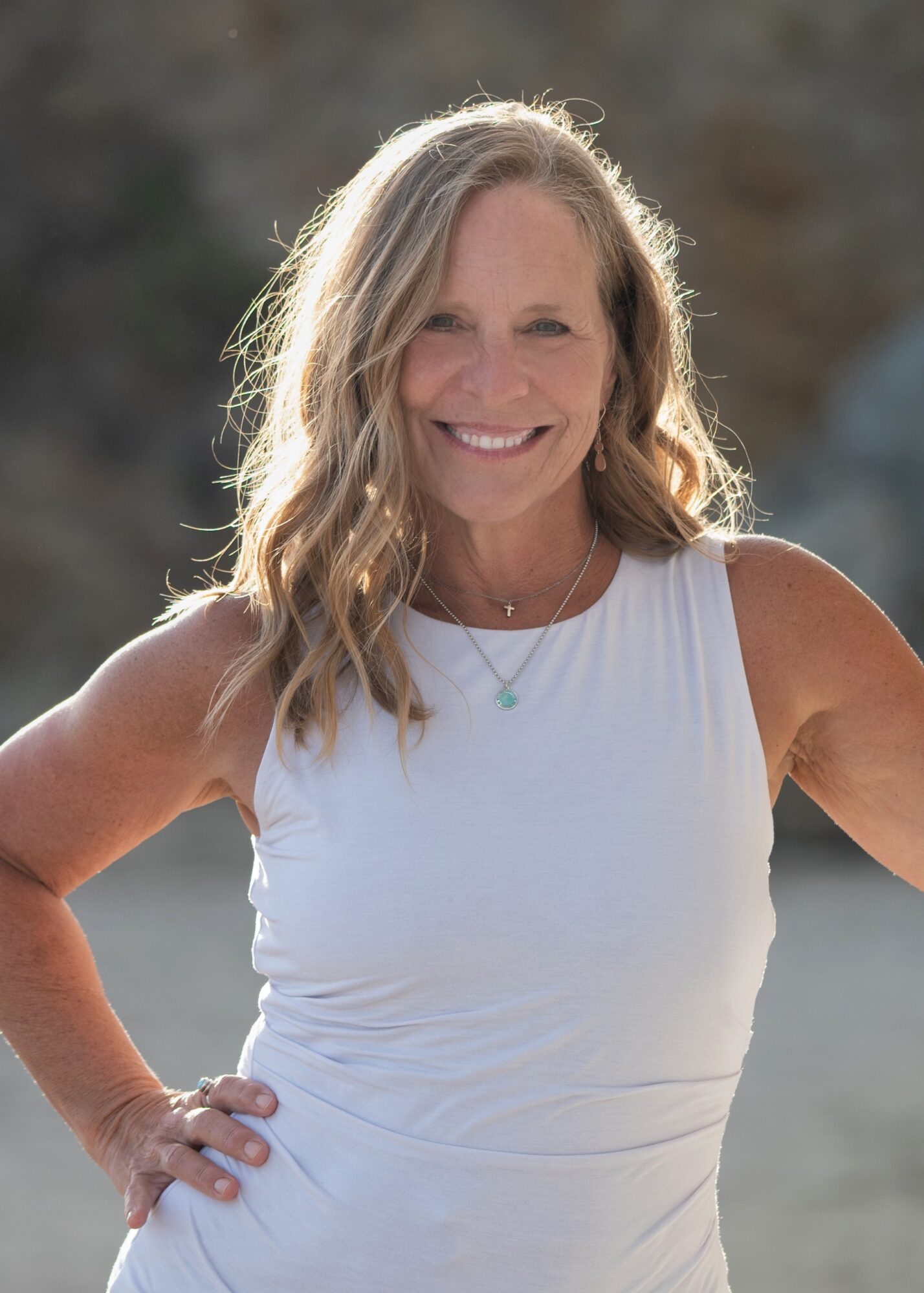 Smiling woman with wavy hair in a white sleeveless top outdoors, sunlight behind her, hand on hip.
