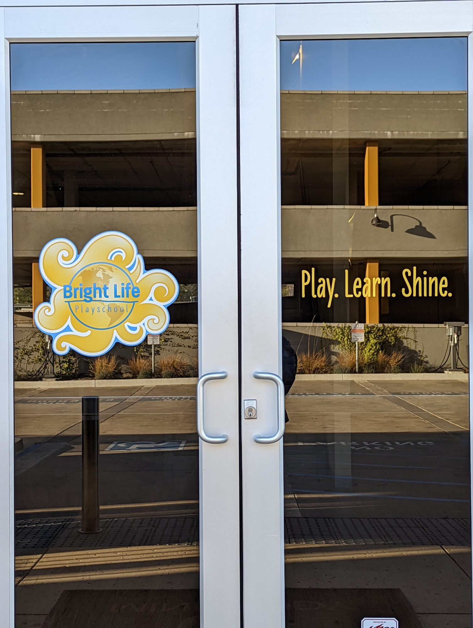 Glass door with signs: Bright Life logo on left, Play. Learn. Shine. on right, reflecting street scene.