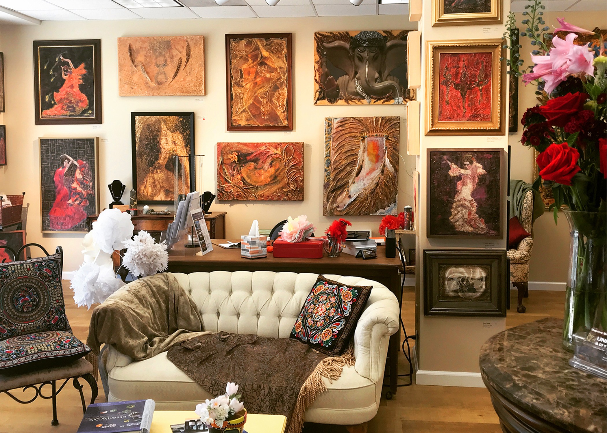Art gallery with framed paintings on walls, a white sofa with pillows, a table with flowers, and a person working at a desk.