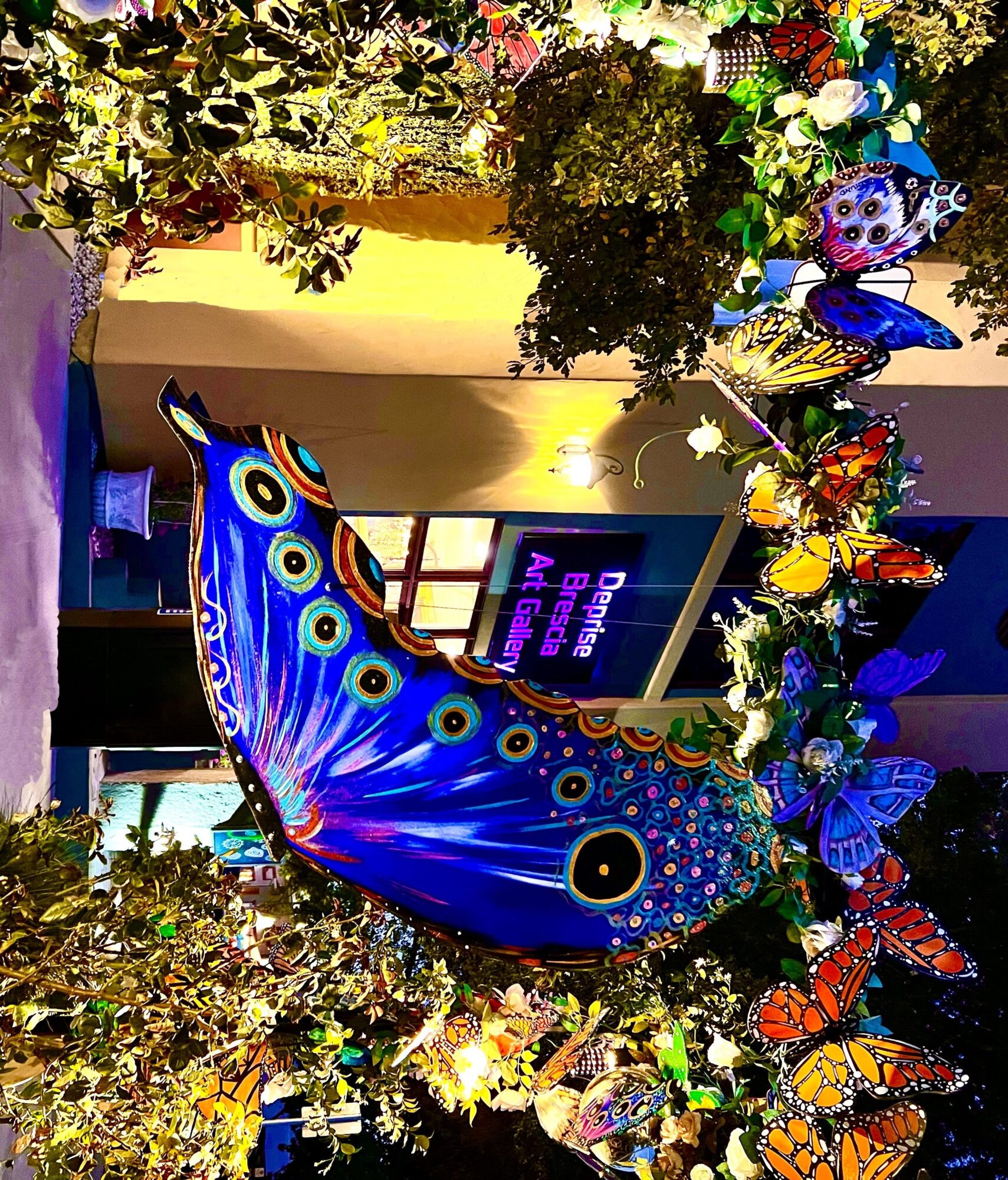 Decorative arch with colorful butterflies and a large peacock feather display, framing a sign for a Brazilian art gallery.
