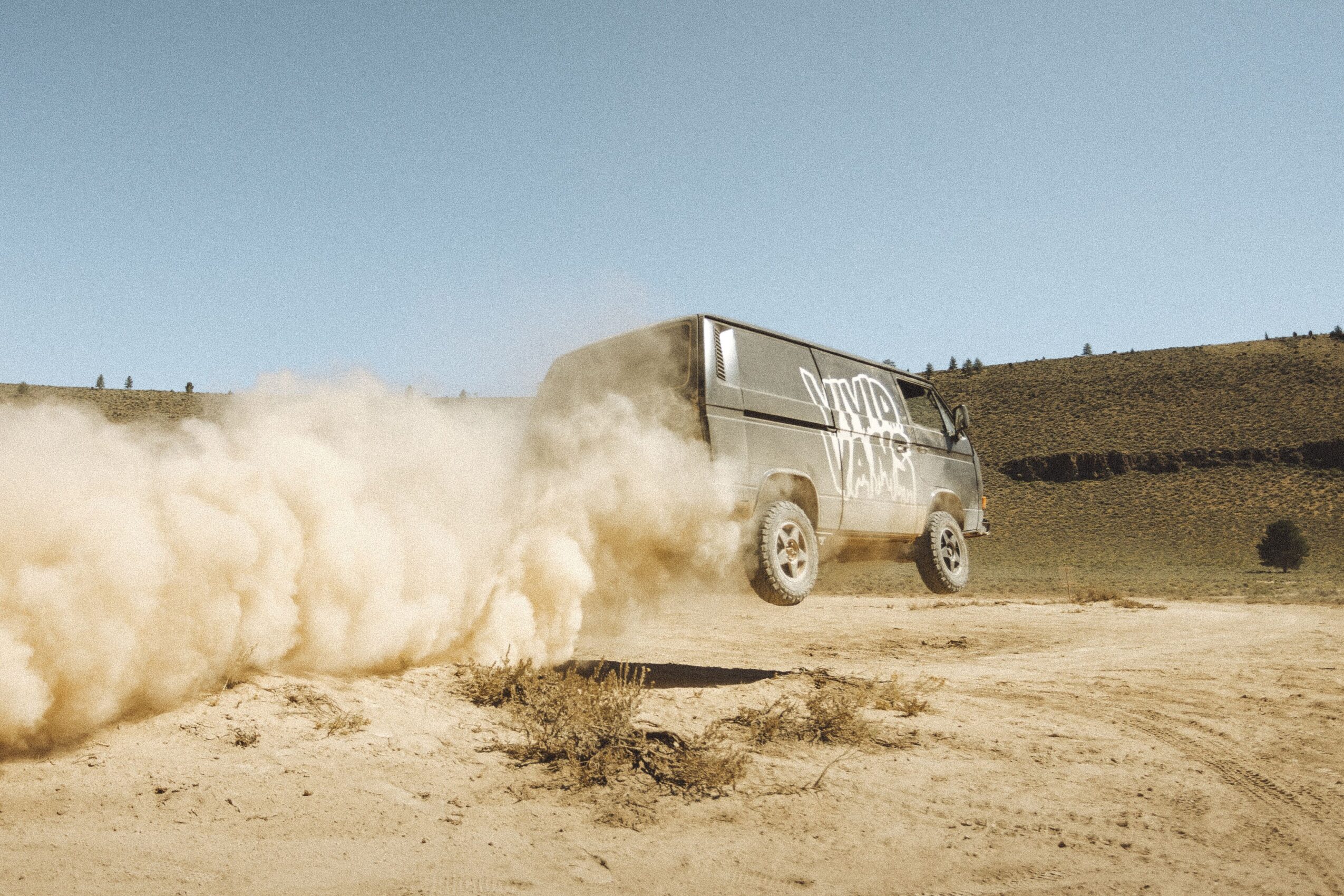 A vehicle kicks up dust as it drives on a dirt road in a rural landscape under a clear sky.