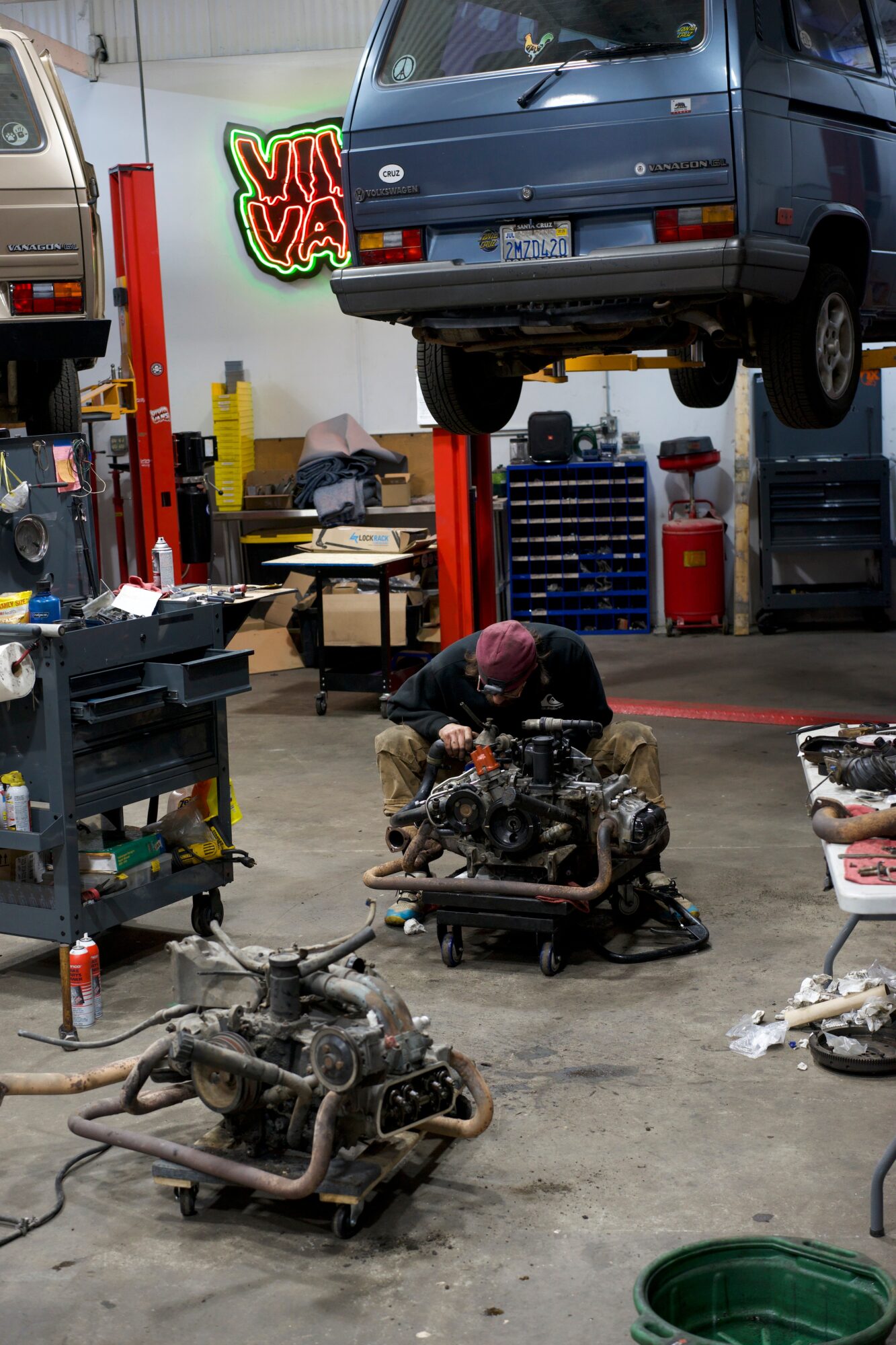 Mechanic working on engine parts in an auto repair shop with a vehicle lifted overhead, tools, and equipment around.