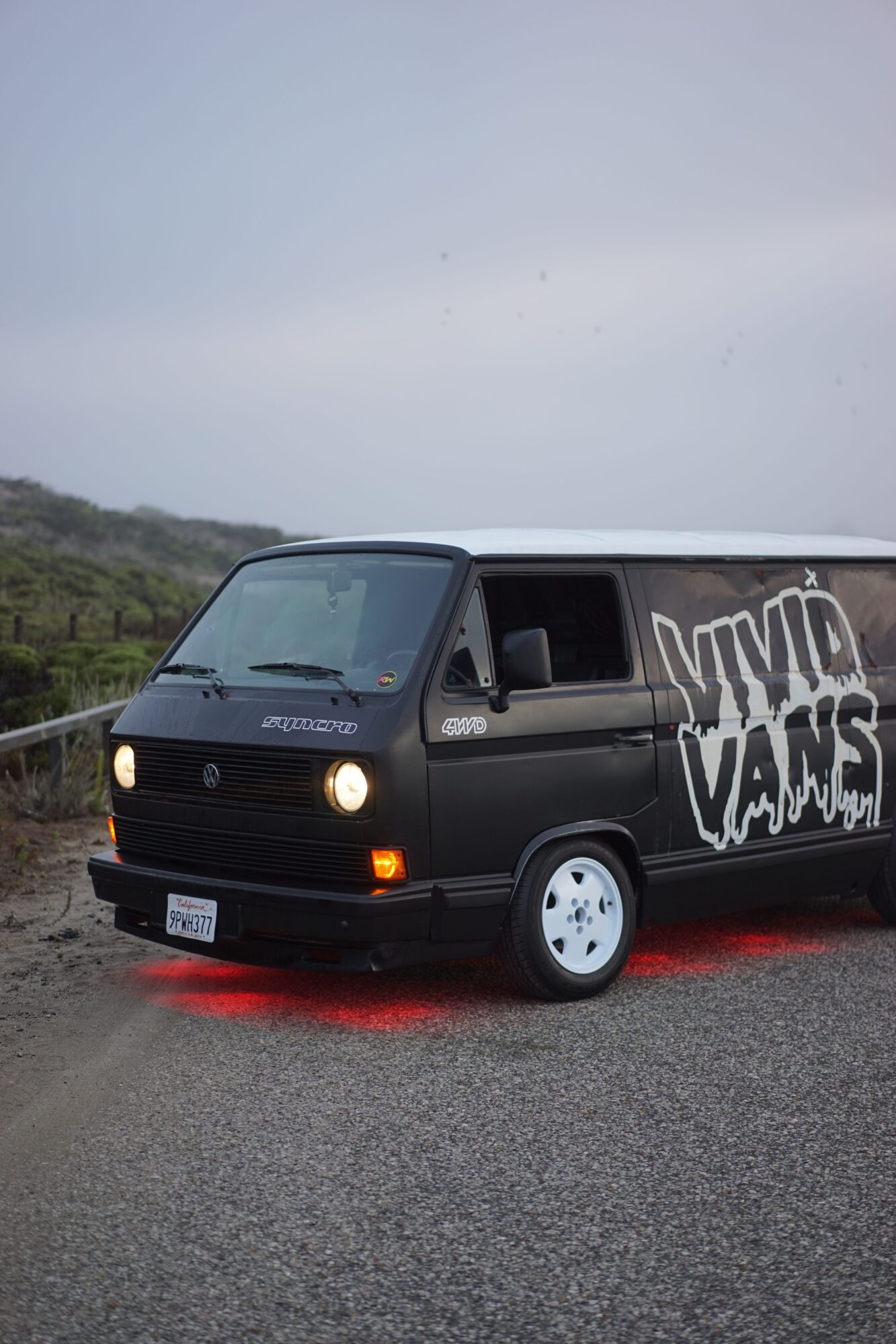 Black van with white graffiti parked on the side of a road, with a landscape background and overcast sky.