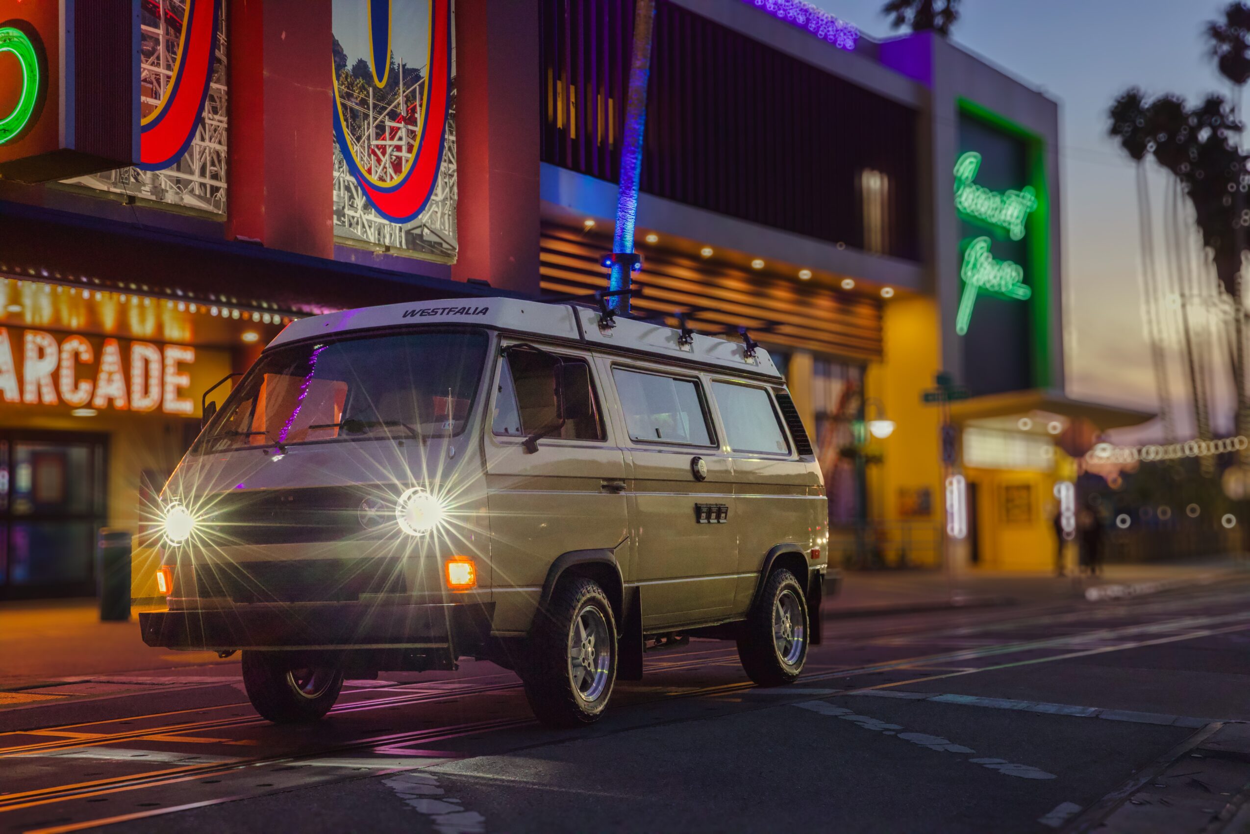 Vintage van parked on street at night with colorful arcade and neon signs in background.