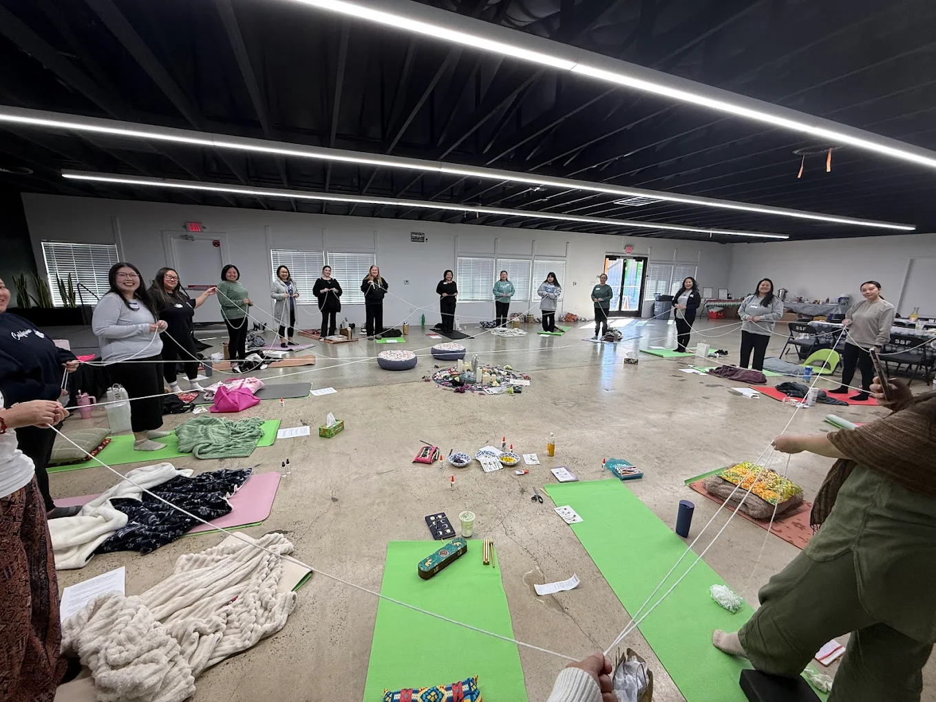 People standing in a circle in a spacious room with black ceiling and bright lighting, participating in a group activity.