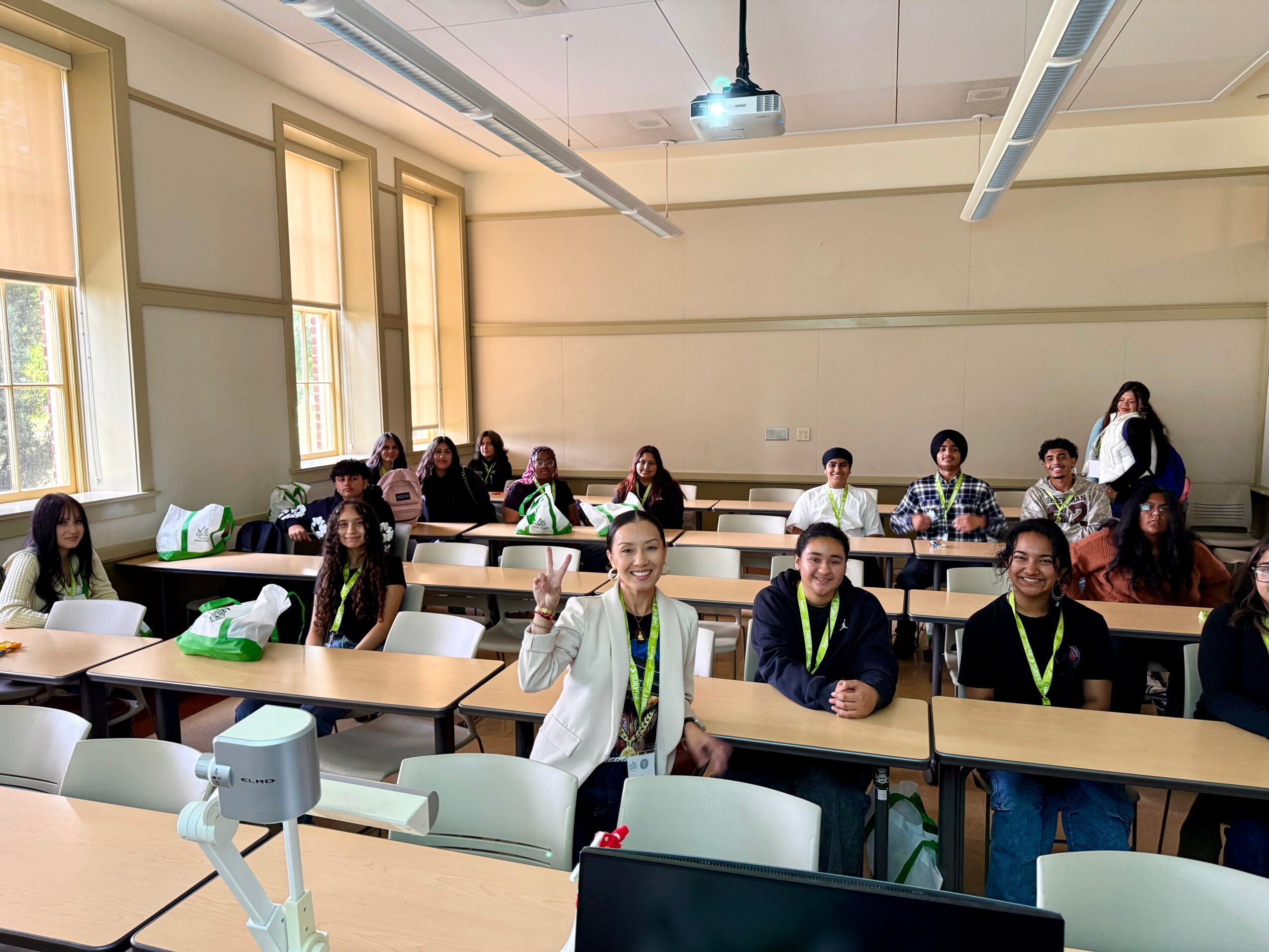 Classroom with students sitting at desks, some smiling and waving, teacher standing at the back, windows on the left.
