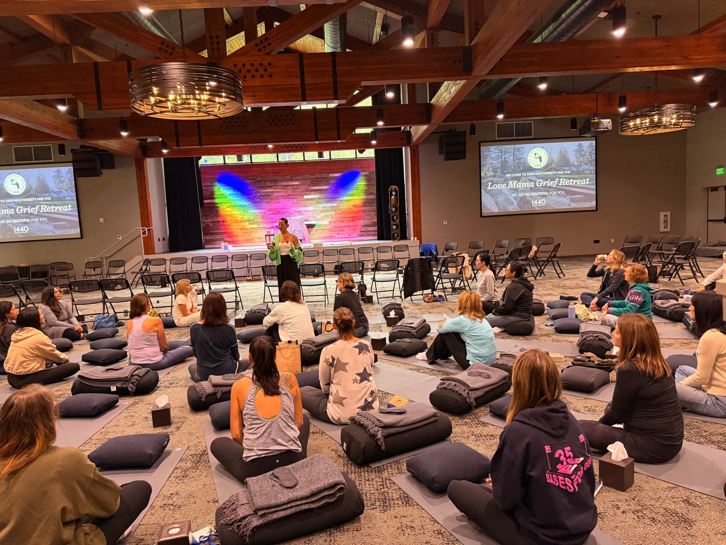 People sitting on cushions facing a stage with a rainbow background, two screens displaying a presentation, in a large room with wooden ceiling beams.
