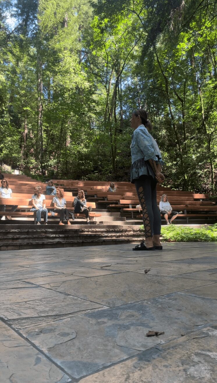 Person standing outdoors near trees and benches, with people sitting on benches in the background, sunlight filtering through leaves.