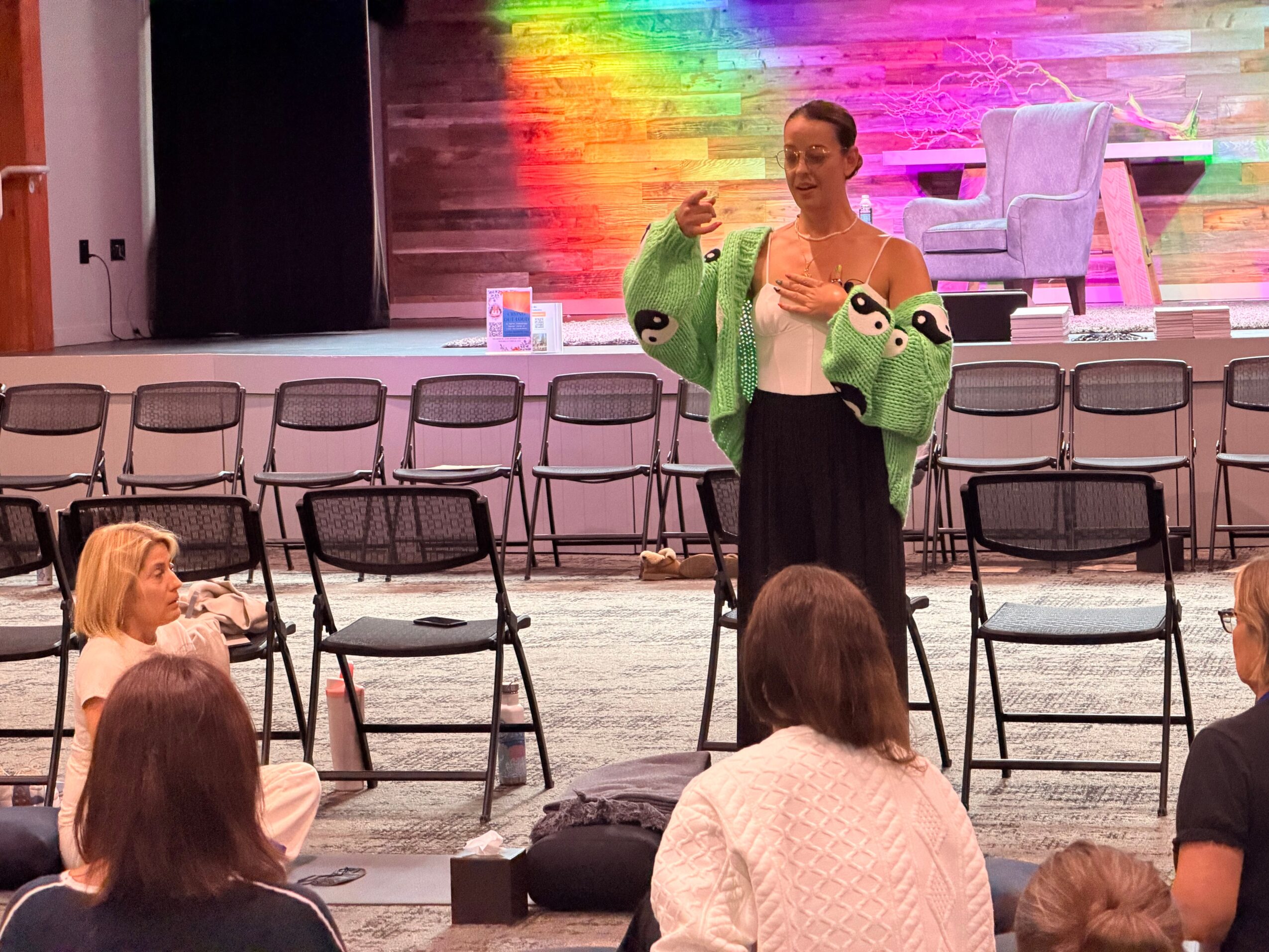 Woman standing on stage with colorful background, wearing a green jacket with animal face designs, speaking to audience.