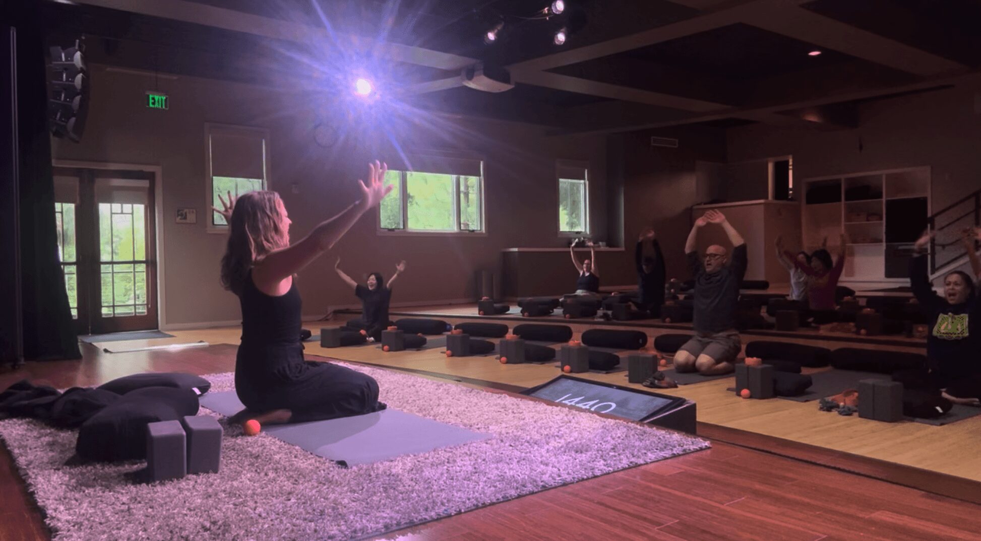 People participating in a yoga or meditation class in a dimly lit room with large windows, mats, and cushions.