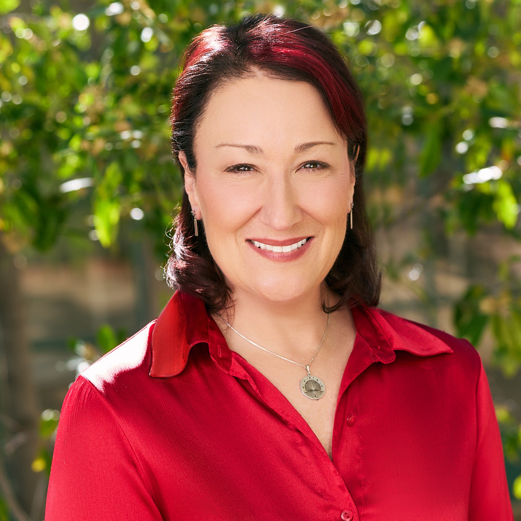 Woman with dark red hair smiling outdoors, wearing a red blouse and silver necklace, greenery in background.