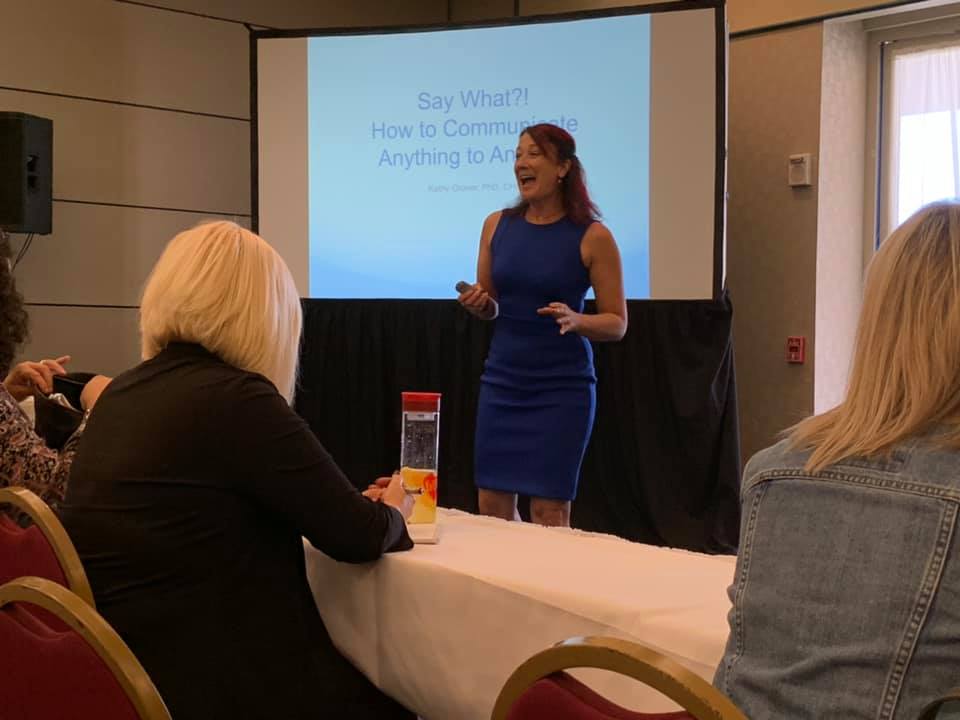 Woman in blue dress giving a presentation in a conference room with audience members seated at a table.