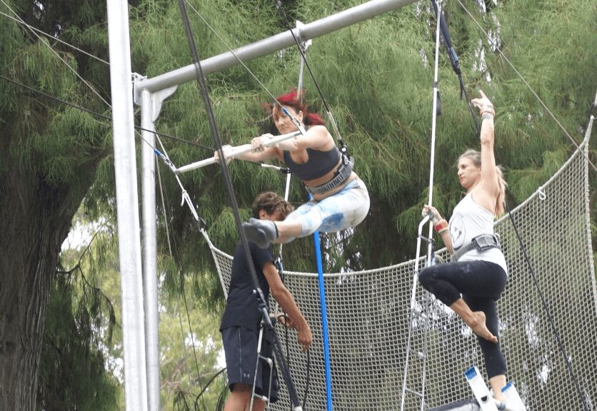 Two women and a man on a trapeze and a trampoline outdoors, surrounded by trees, with safety nets and ropes visible.