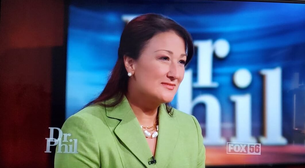 Woman with dark hair in a green blazer on a TV set with blue background and text