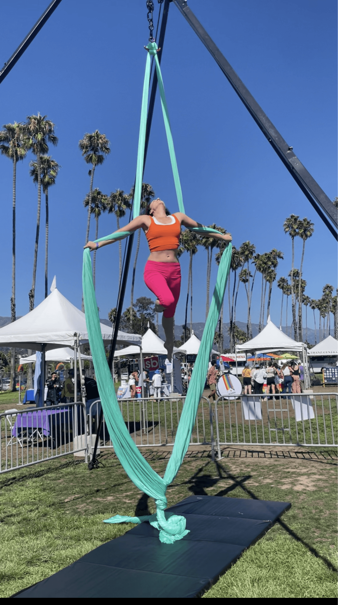 A person performing aerial silk acrobatics outdoors with palm trees and tents in background.