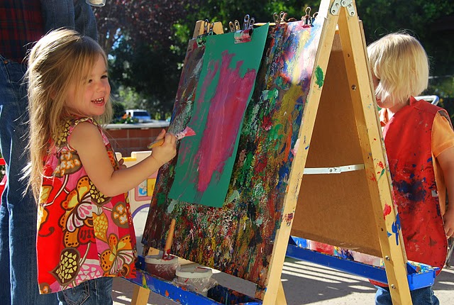 Children painting on outdoor easels with colorful artwork, surrounded by trees and sunlight.