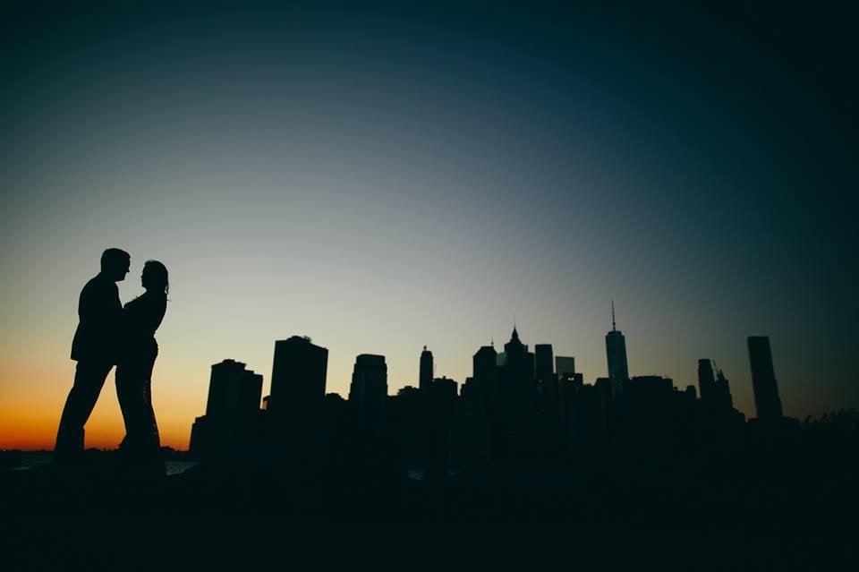 Silhouette of a couple standing close together against a city skyline at sunset or dusk, with a darkening sky.