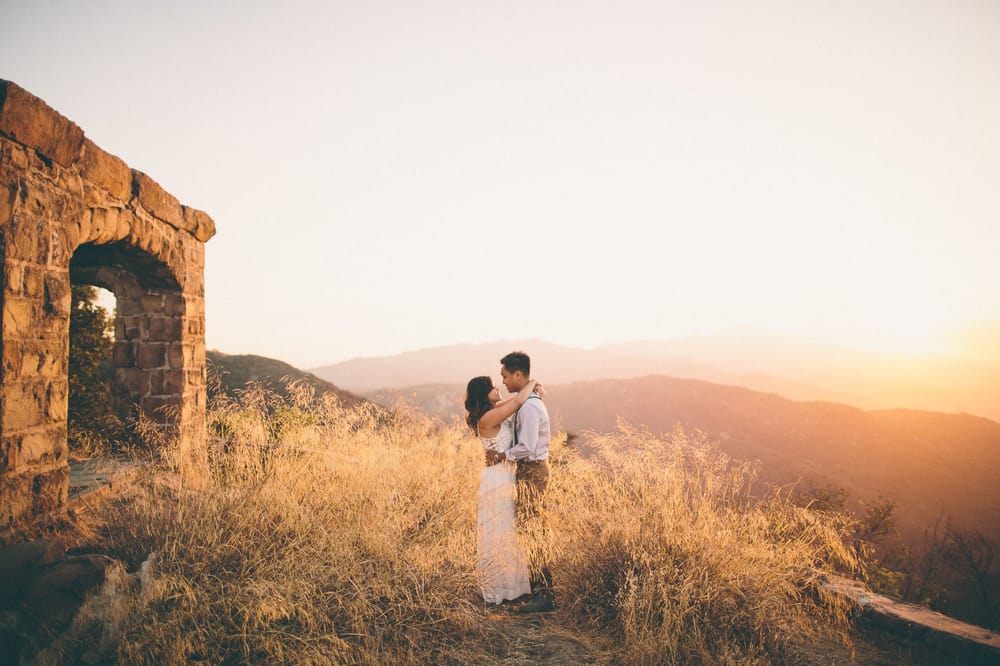A couple stands close together in a grassy field during sunset, with a stone structure on the left and mountains in the background.
