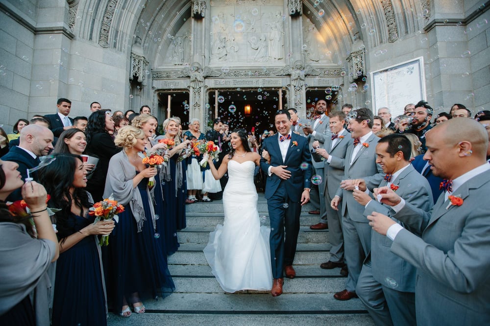 Bride and groom walking down steps surrounded by guests celebrating outside a church, holding flowers and smiling.