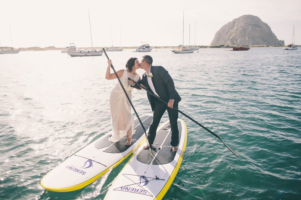 Two people in formal attire stand on a paddleboard, leaning in for a kiss on the water with boats and a island in background.