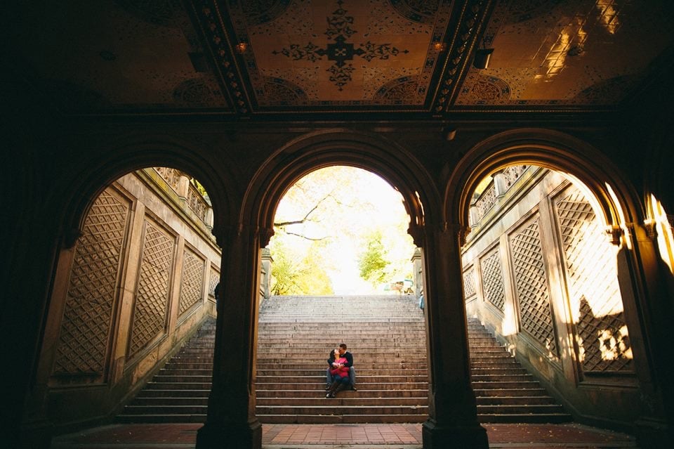 Person sitting on stairs outside, viewed through an ornate archway with columns, steps leading upward, and surrounding architecture.