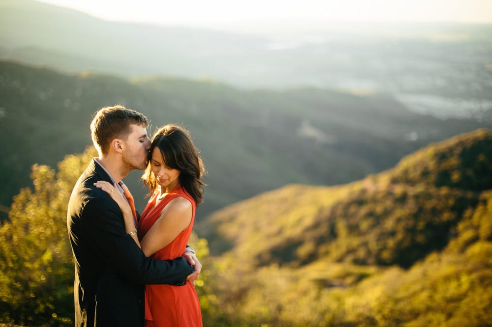 A couple embraces outdoors with a scenic landscape of rolling hills and a distant horizon in the background.