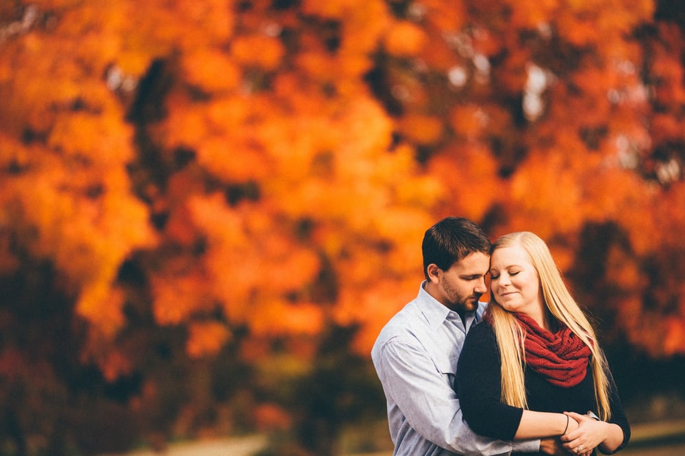 A man and woman stand close together outdoors with autumn-colored trees in the background.