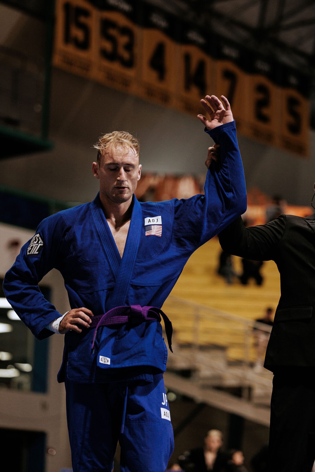 Judo athlete in blue gi with purple belt raising hand, in an indoor sports arena with banners and spectators.