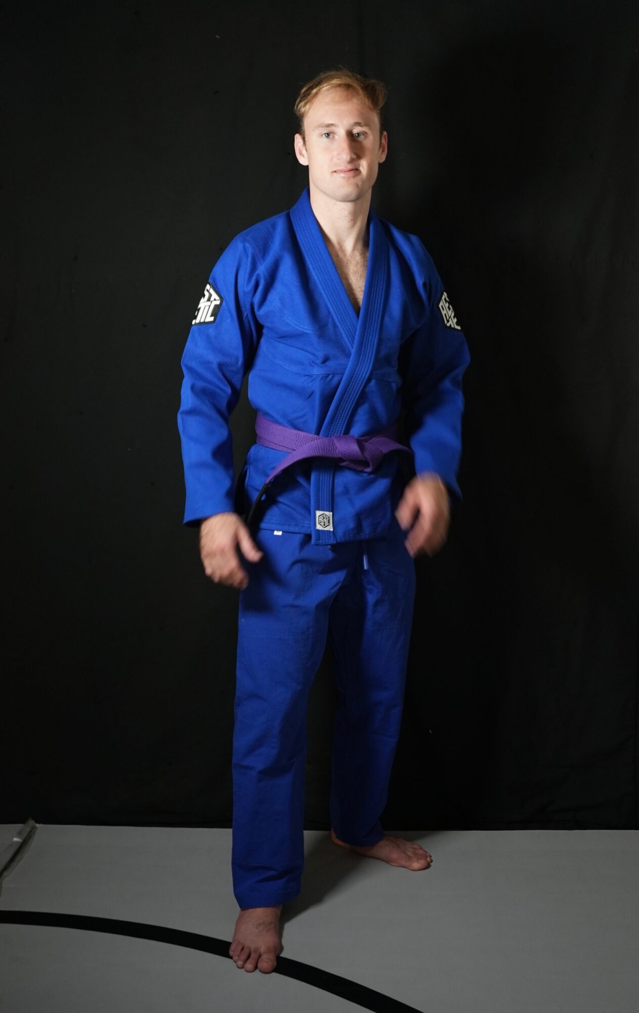 Person standing in a blue martial arts gi with a purple belt, facing forward against a black background.