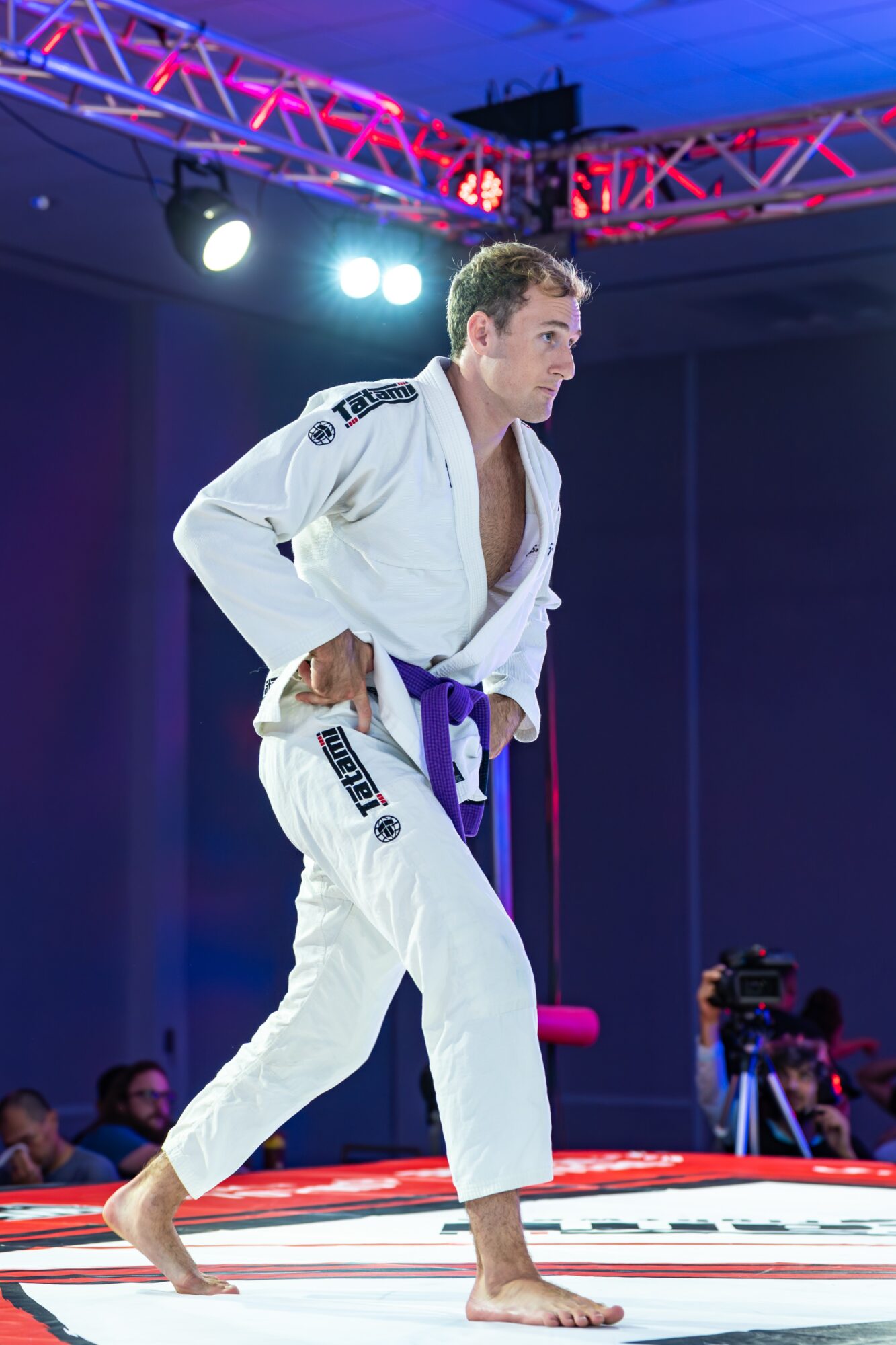 Martial artist in a white gi with a purple belt standing on a mat, with stage lights and audience in background.