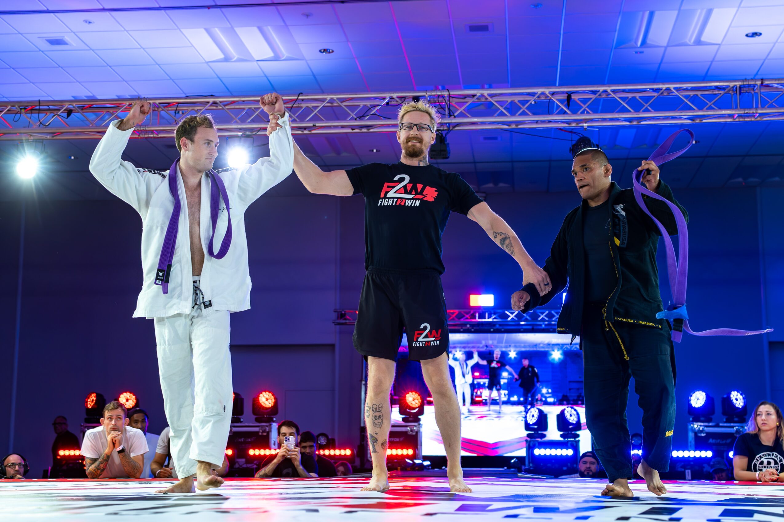 Three men on stage, one in a martial arts uniform, holding hands, with a blue-lit background and audience.