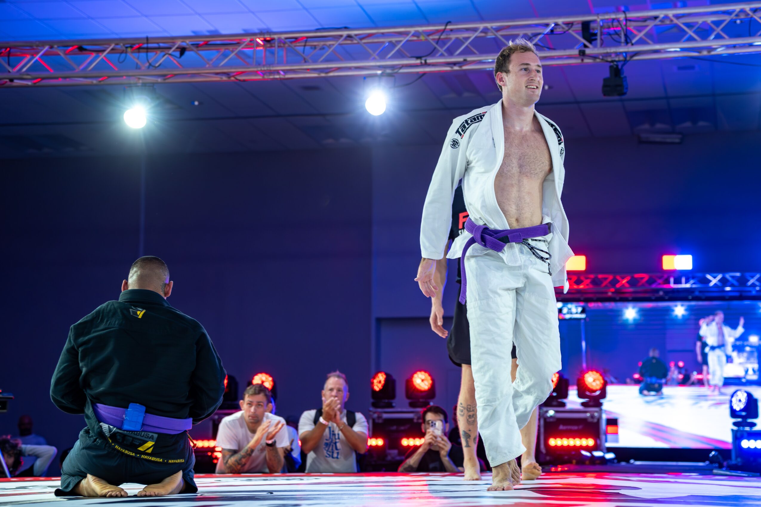 Martial artist in a white gi with a purple belt standing on stage, audience clapping, bright lights, and a large screen in background.