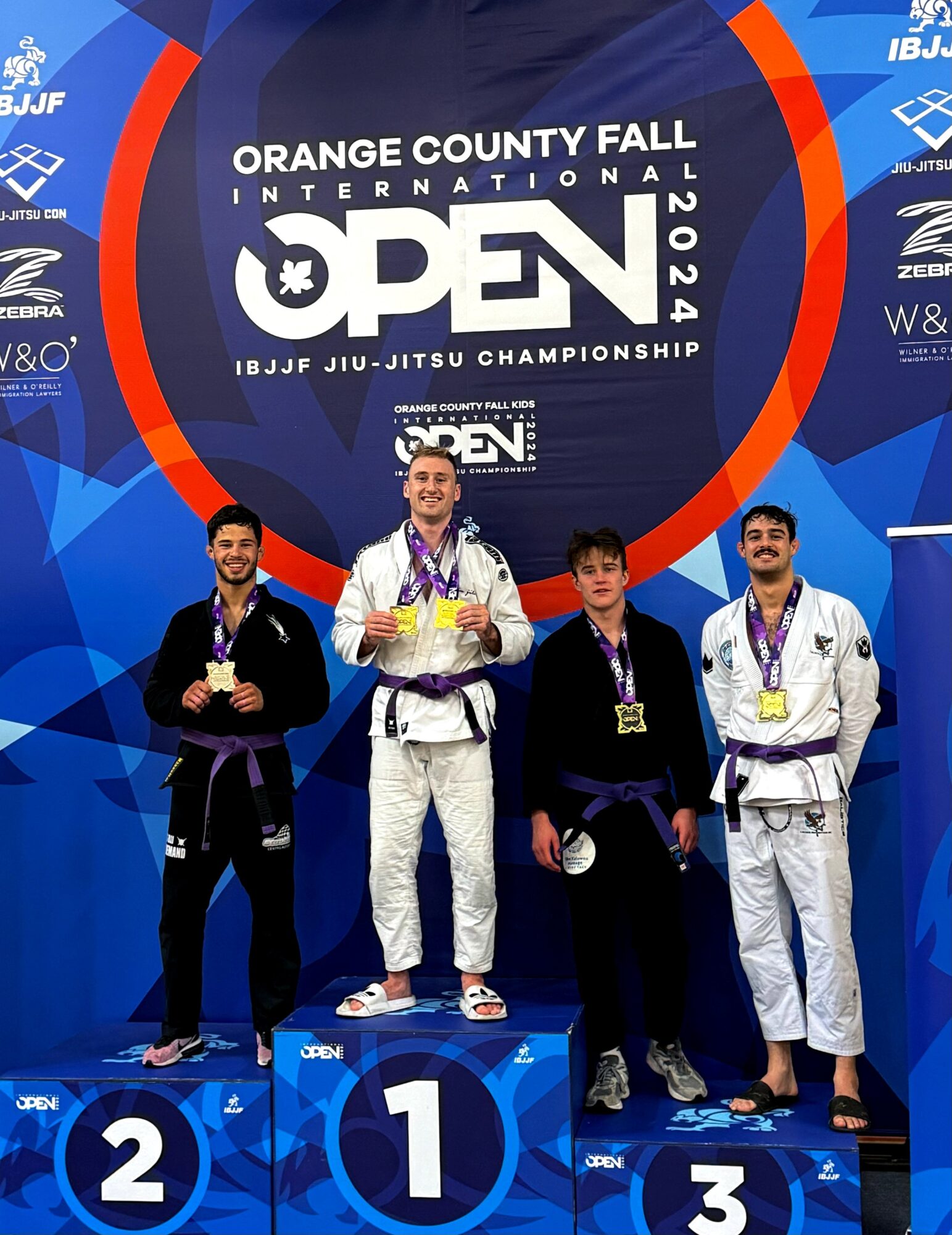 Four athletes on a podium with medals at the IBJJF Jiu-Jitsu Championship, first place in the center, others on sides, backdrop with event logo.