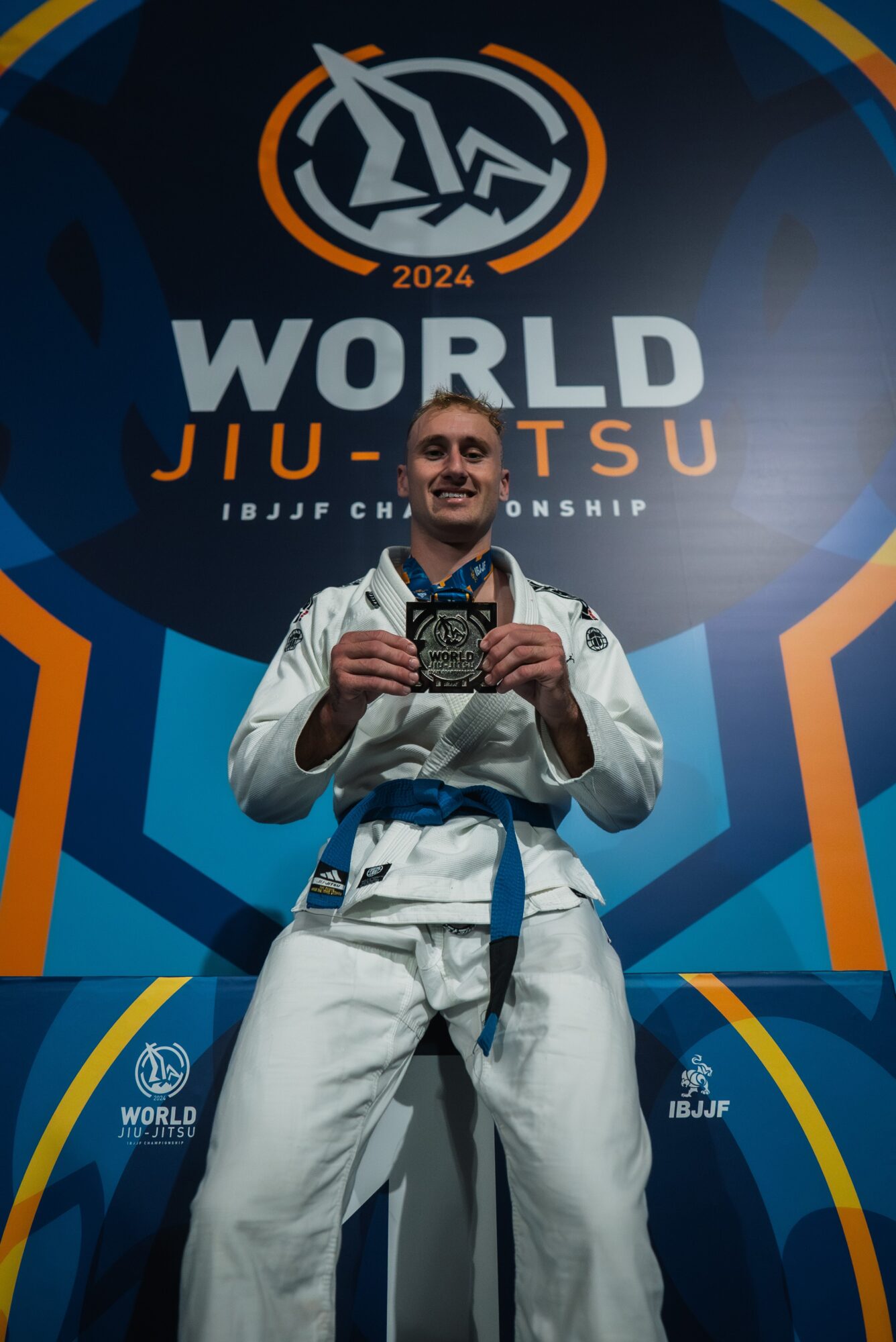 Person in a white martial arts uniform holding a medal, standing in front of a World Jiu-Jitsu Championship backdrop.