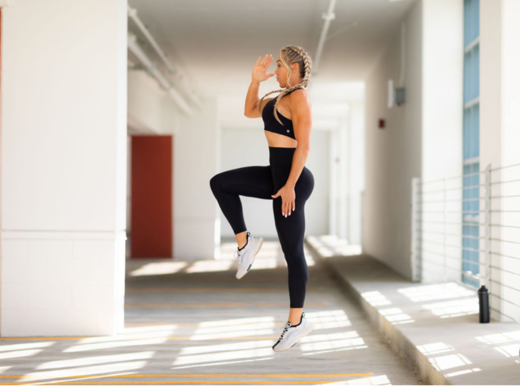Woman in athletic wear jumps in a bright, modern indoor space with large windows and sunlight streaming in.