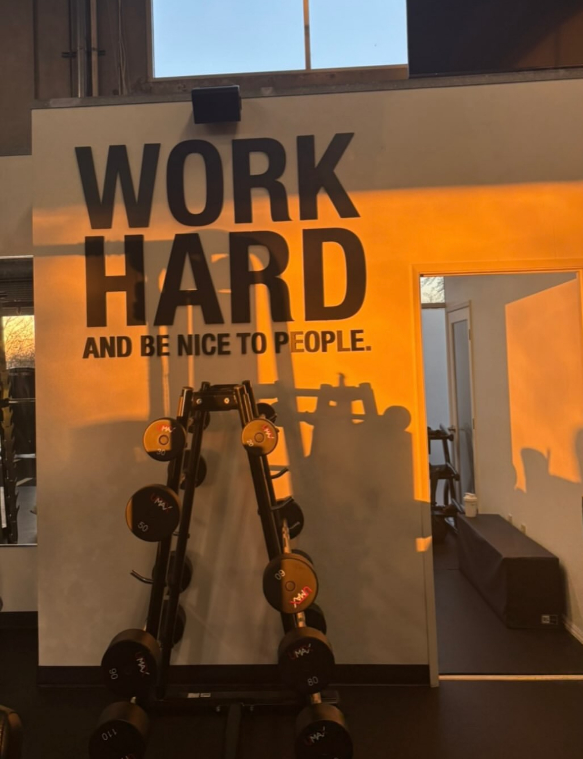 Gym wall with motivational sign and exercise equipment, sunlight casting shadows, window in background.