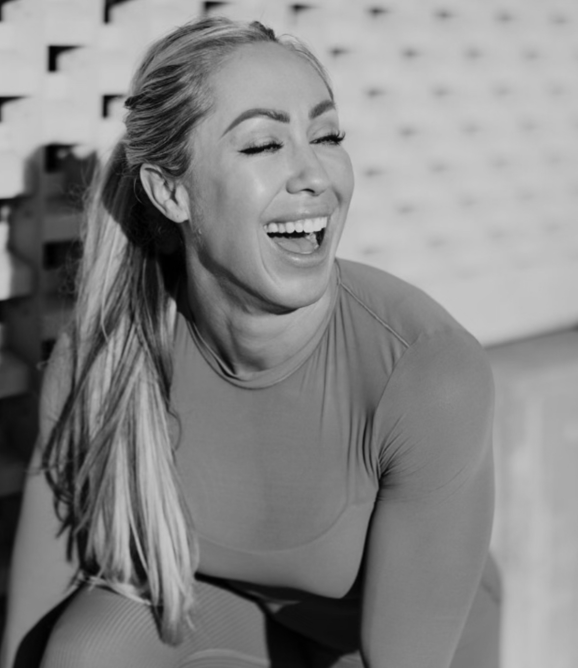 Woman laughing with eyes closed, long hair, wearing a fitted top, in black and white, smiling broadly.