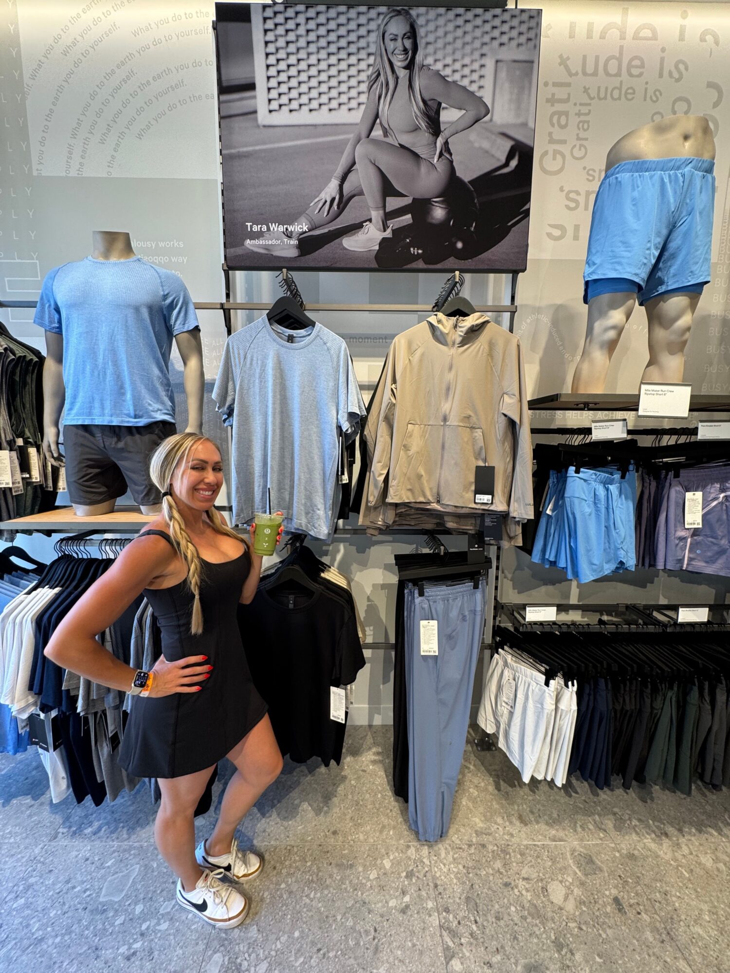 Two women stand in a clothing store, one holding a drink, surrounded by racks of clothes and a large black-and-white photo.
