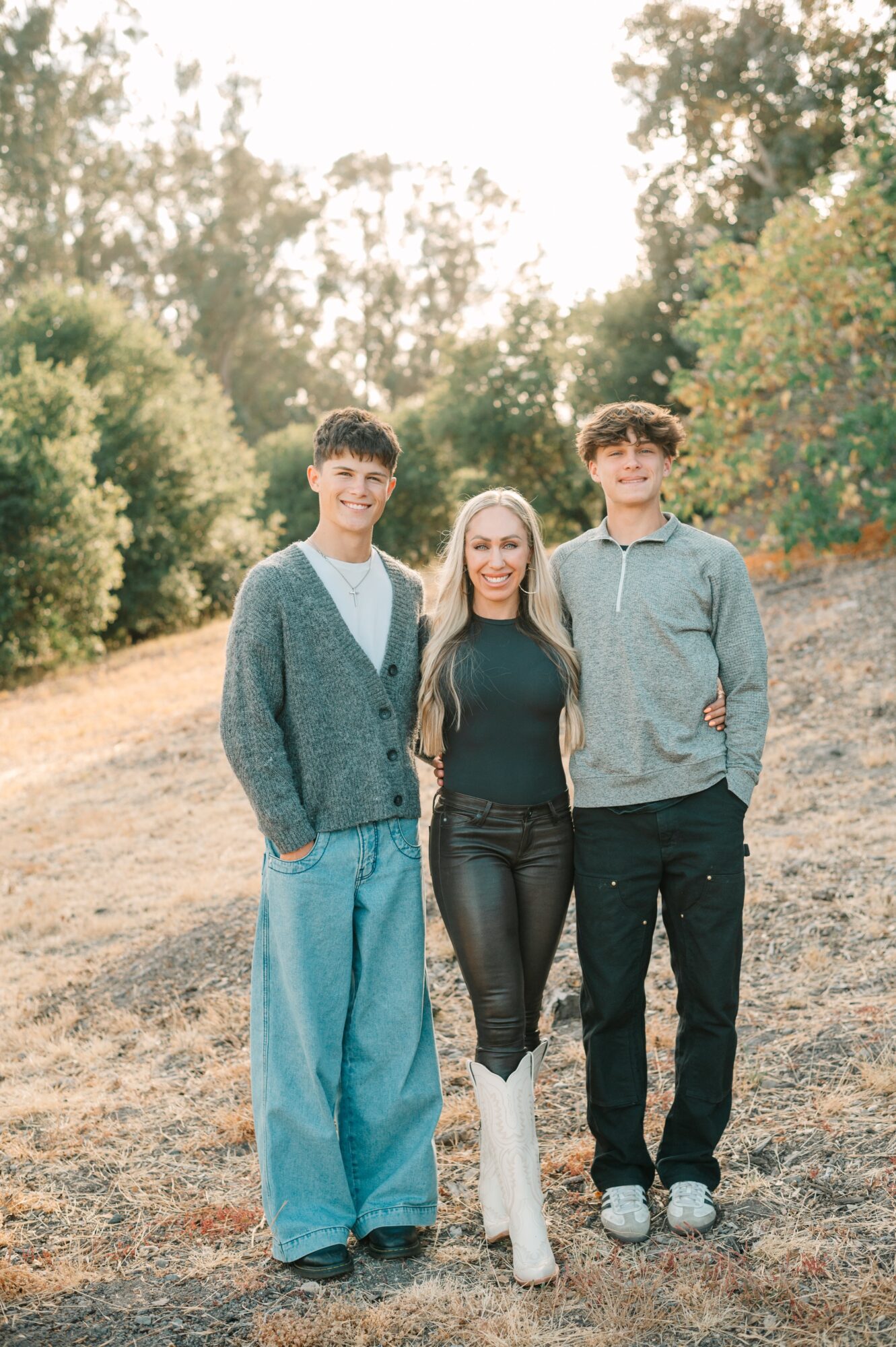 Three people standing outdoors on a grassy hill with trees in the background, smiling at the camera.