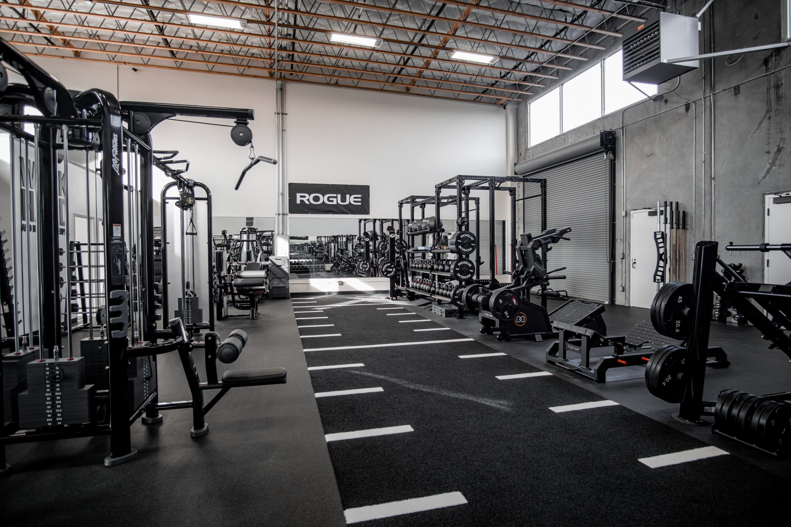 Empty gym with weightlifting equipment, benches, and a Rogue sign on the wall, illuminated by natural light from windows.