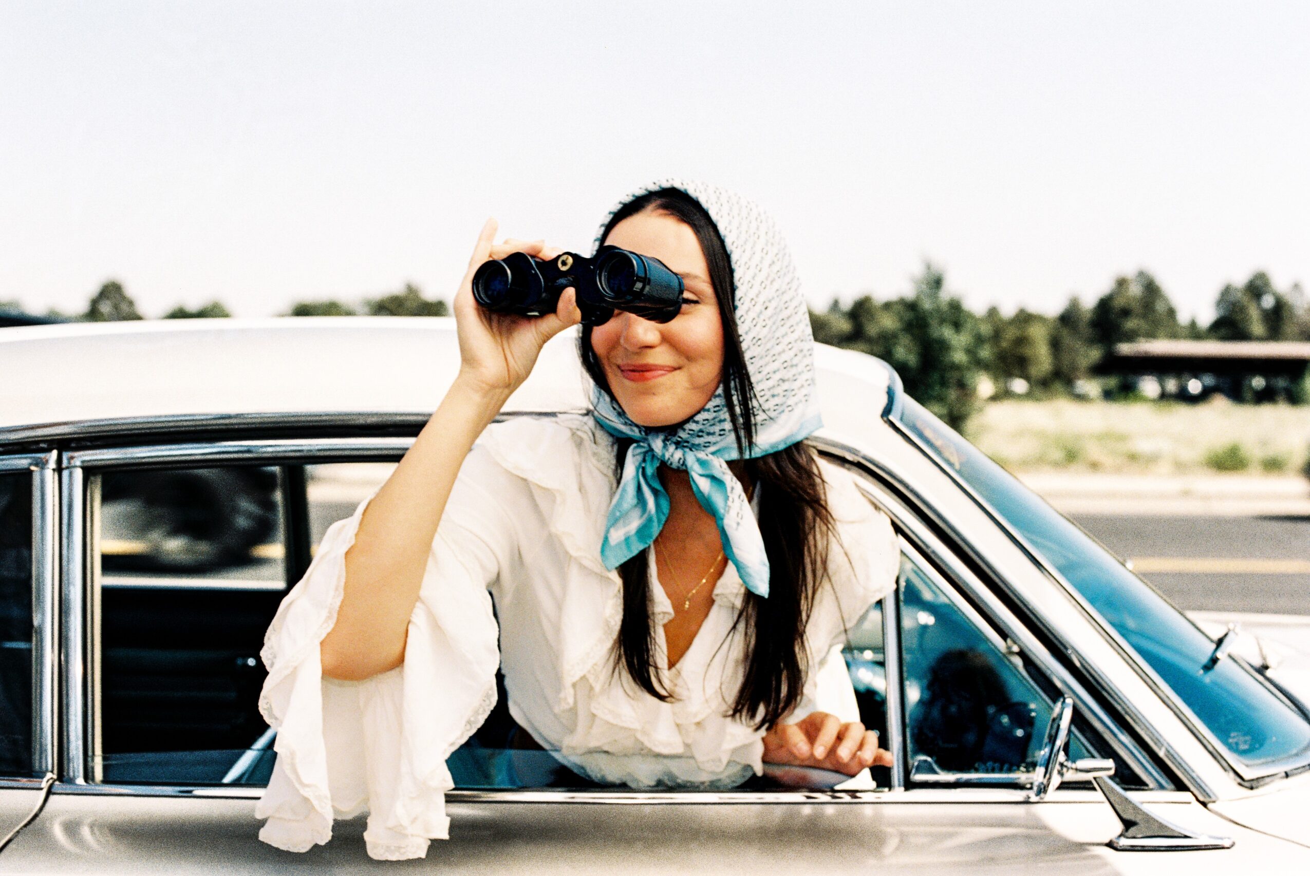 Woman with a headscarf looking through binoculars from car window, outdoors, sunny day.