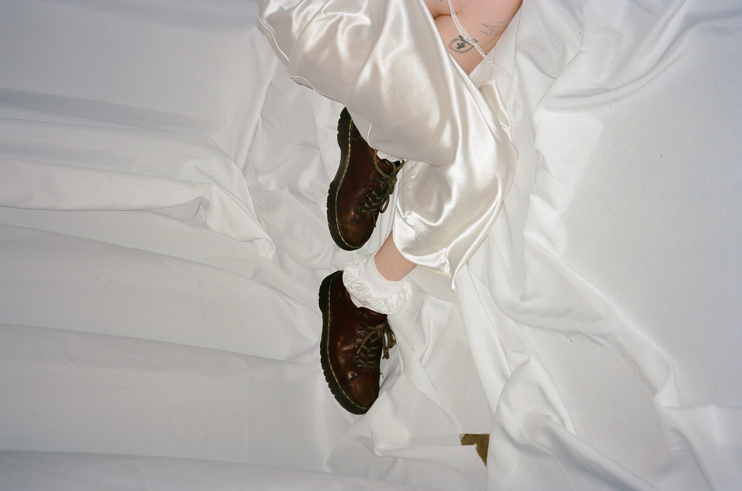 Person wearing white satin pajamas and brown shoes lying on white bed sheets, legs crossed.