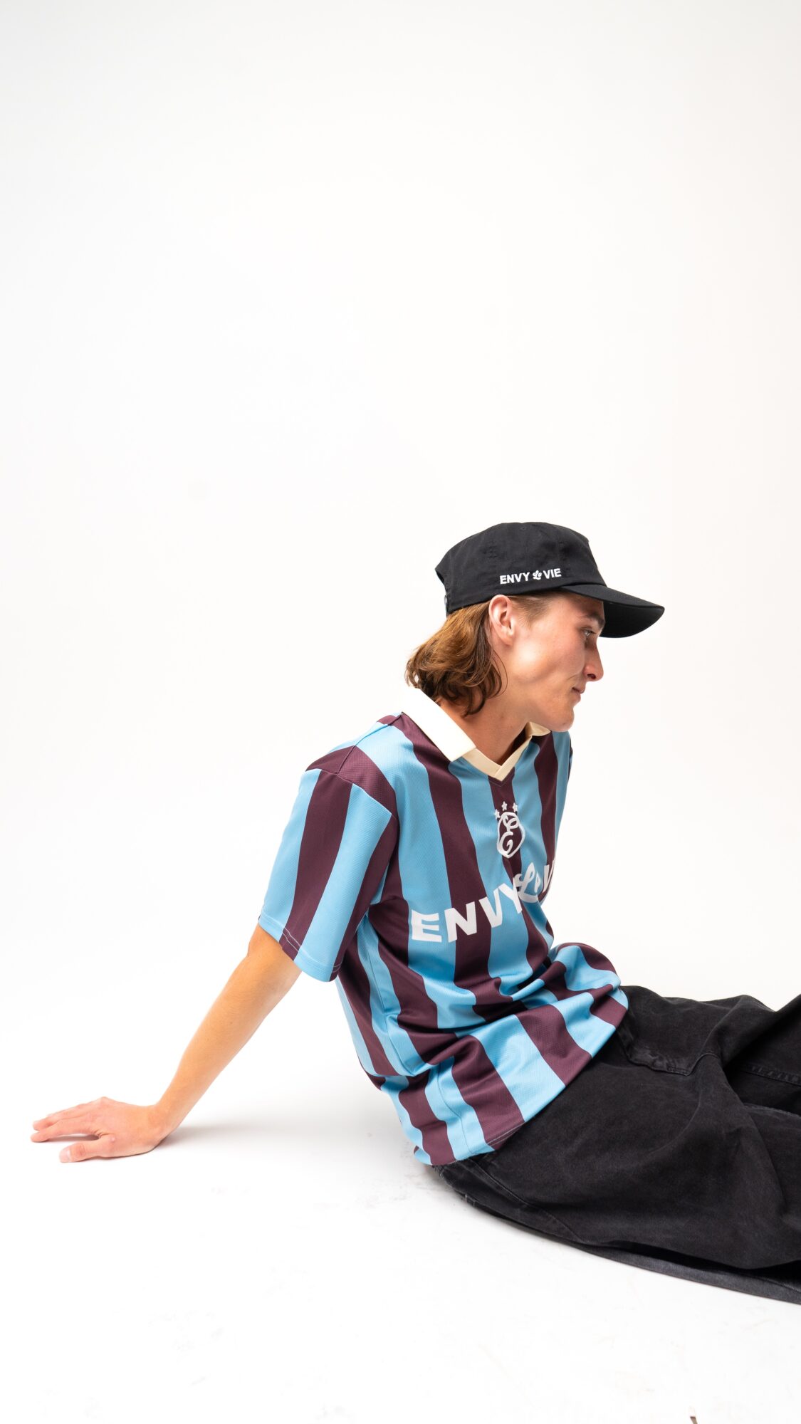 Person sitting on the floor wearing a striped shirt, black cap, and black pants, facing right, with a plain white background.
