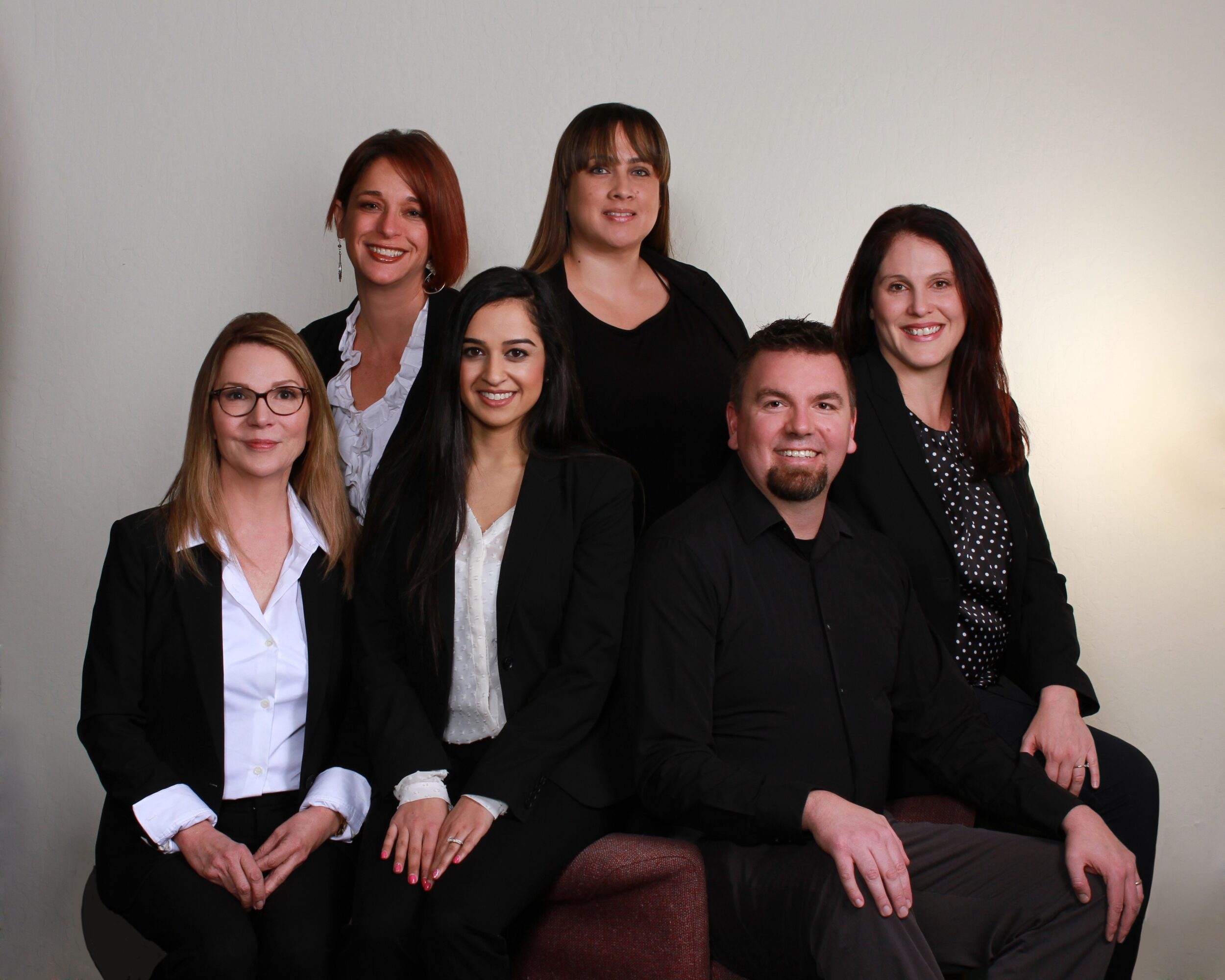 Group of six people, five women and one man, posing together against a plain background, dressed in business attire.