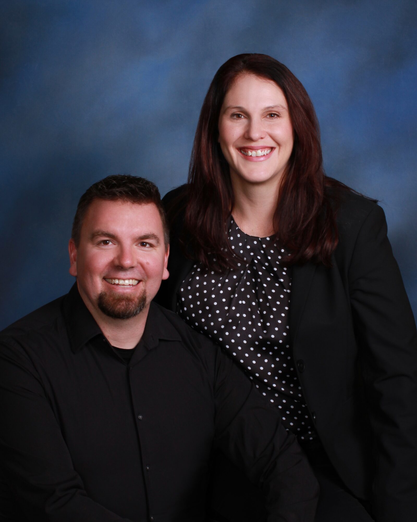 A smiling man and woman pose together against a blue background, both dressed in dark clothing. The woman has long dark hair.