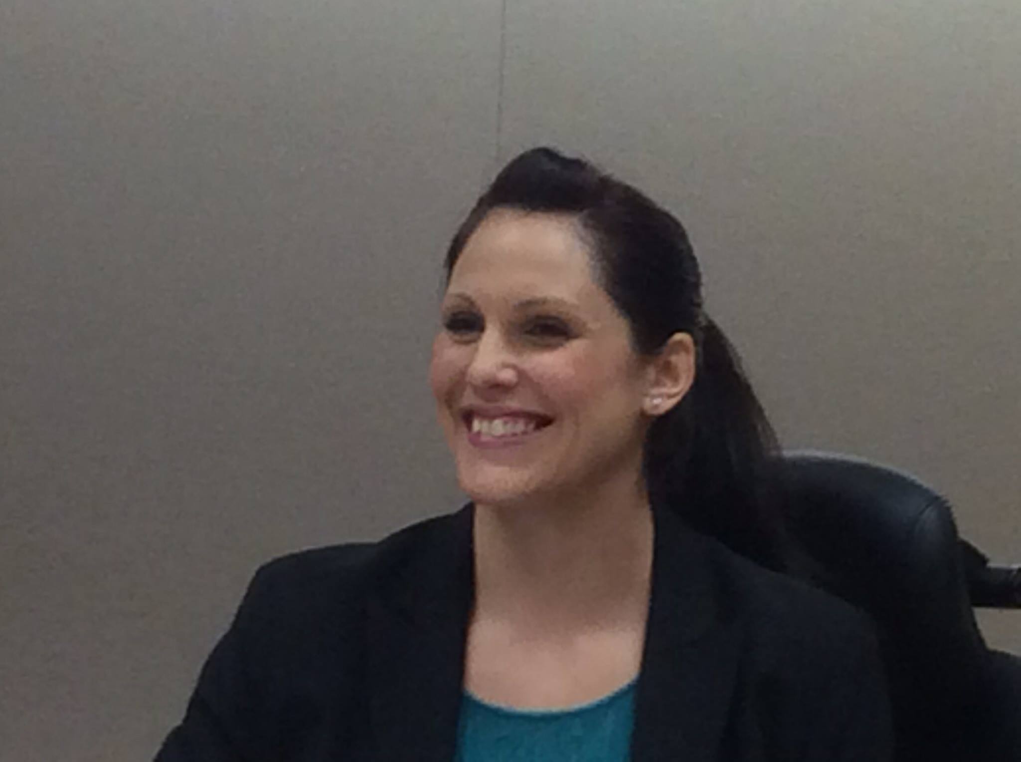 Smiling woman with dark hair in a black blazer and teal top, sitting in a chair against a plain background.