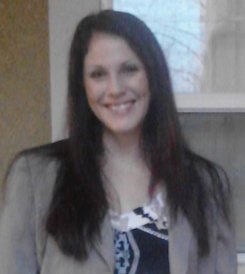 Smiling woman with long dark hair, wearing a light-colored blazer and patterned top, standing indoors near a window.