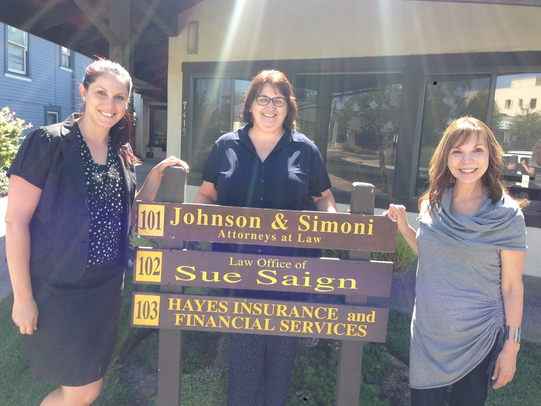 Three women standing outdoors behind a sign with law office names, sunlight shining on them, smiling faces.