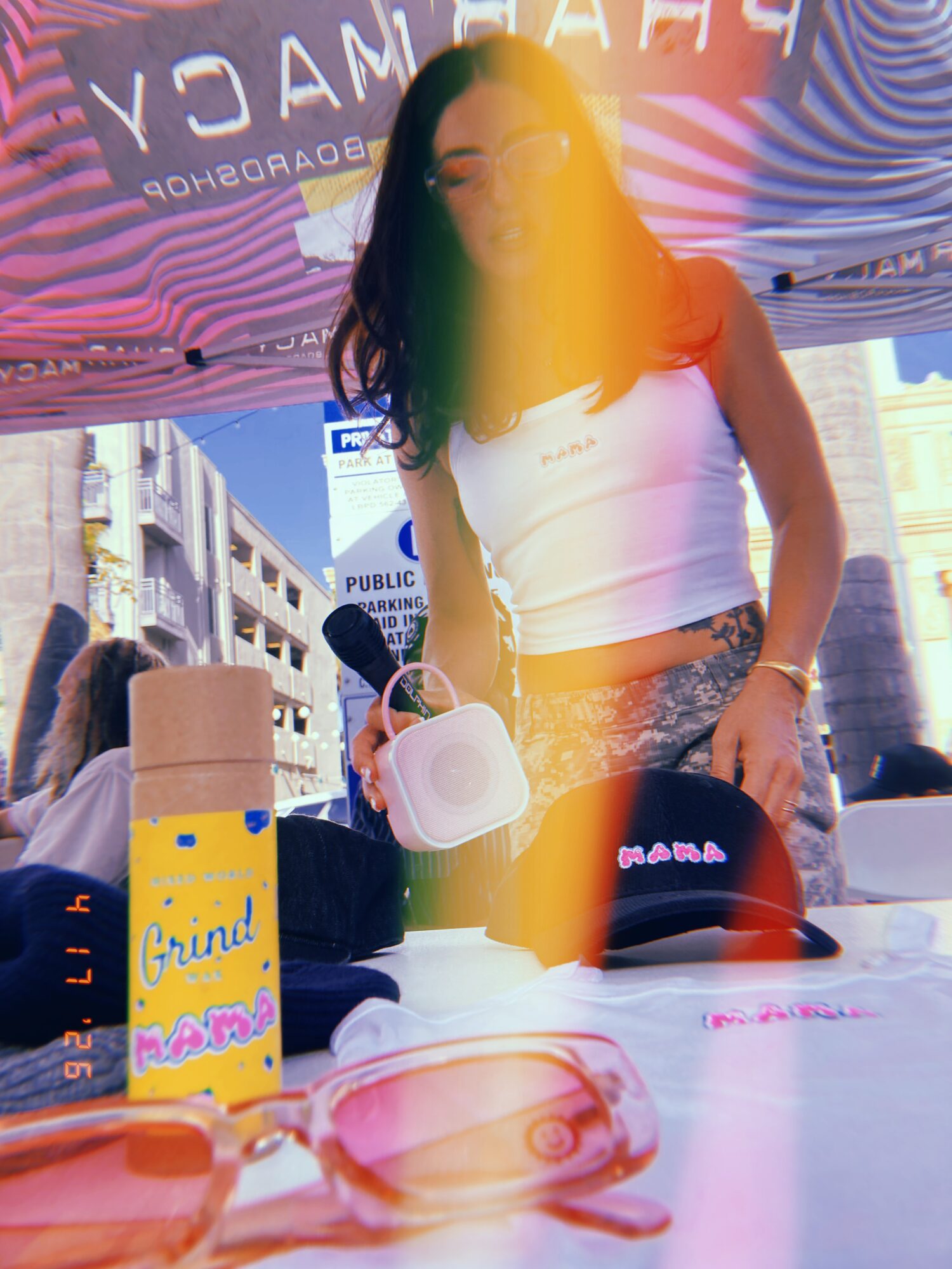 Young woman standing at an outdoor event table with drinks and items, wearing a white crop top and patterned pants.