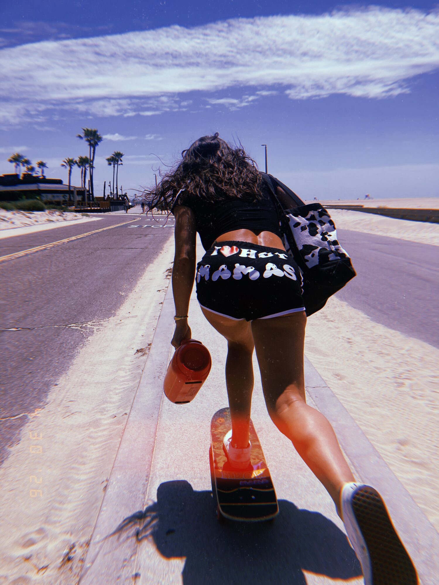 Person skateboarding on a beachside road with palm trees and blue sky, carrying a bag and holding a hat, facing away.